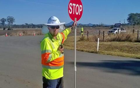 Beau standing beside a road wearing hi vis shirt and safety hat. He is holding a stop sign out beside him